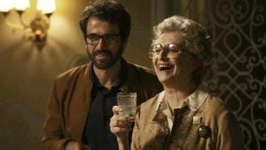 em um cenário de novela "O Outro Lado do Paraíso", um homem branco barbudo e usando óculos está em pé ao lado de uma mulher mais velha sorridente