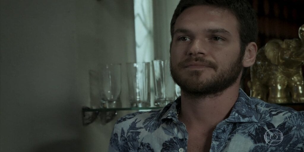 Rubinho da novela A Força do Querer olha para o lado esquerdo com expressão séria usando uma camisa branca com flores azuis