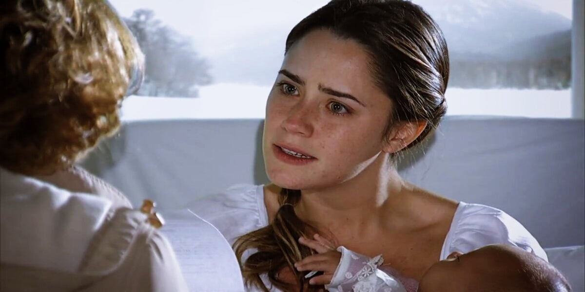 Ana em A Vida da Gente está deitada em cama de hospital segurando uma bebê