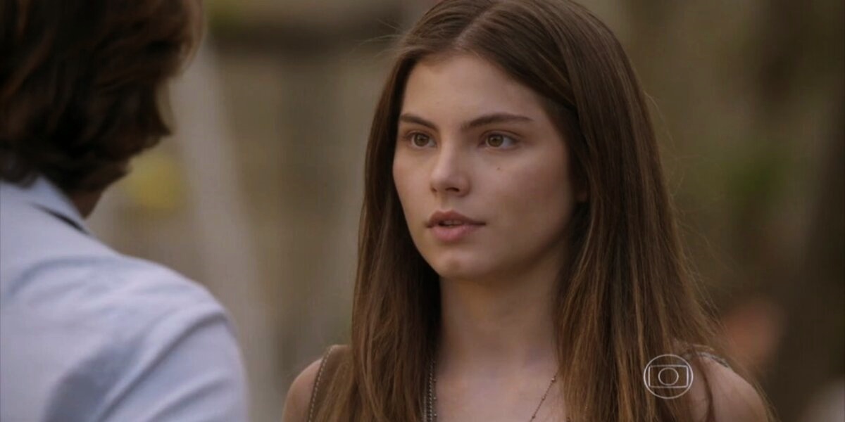 Bianca, jovem menina branca de cabelos longos, lisos e castanhos está em cena da novela Malhação olhando para um homem de costas usando camisa azul clara