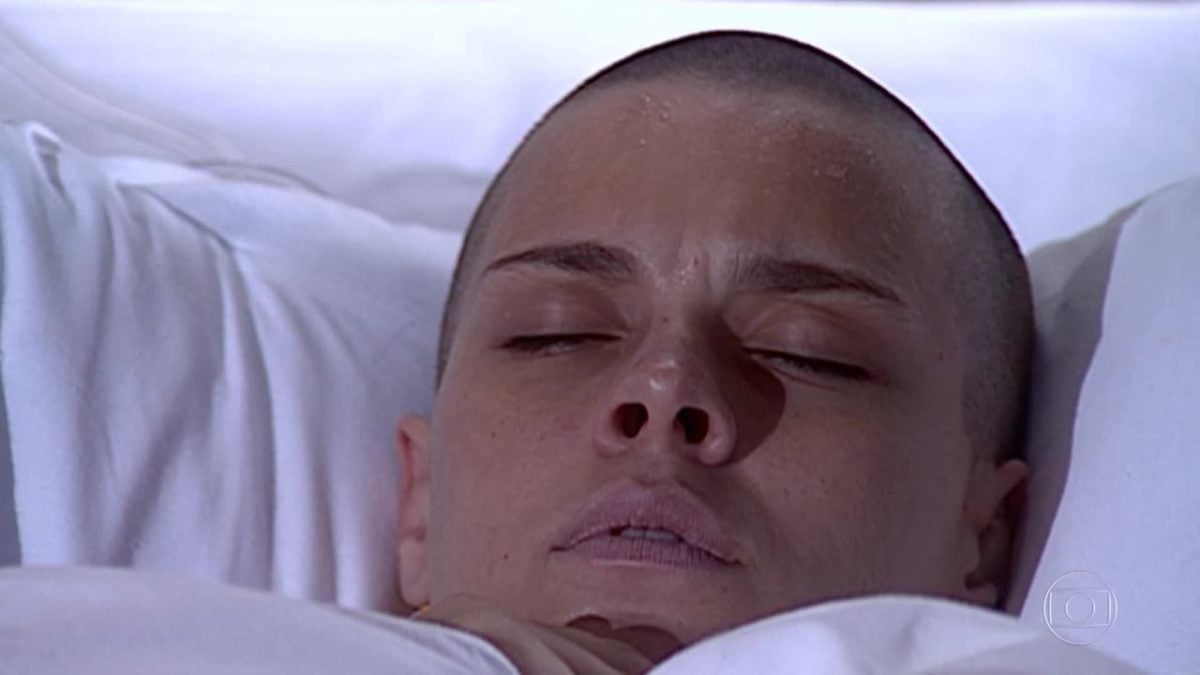 Camila, mulher branca e careca, está deitada com os olhos fechados em cena da novela Laços de Família