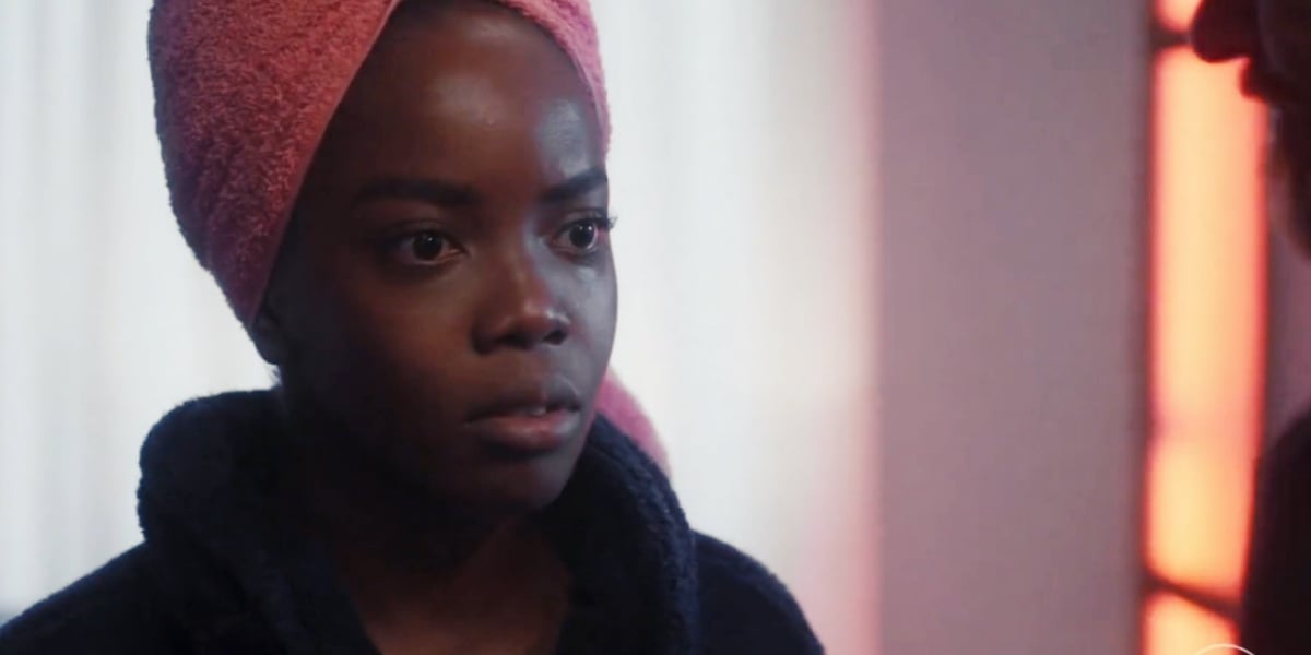 Em Vai na Fé, Kate (Clara Moneke) usa roupão azul escuro, toalha rosa amarrada na cabeça e demonstra apreensão