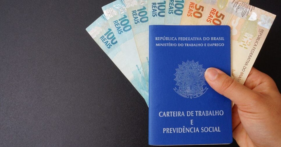 Pessoa segurando carteira de trabalho e notas de real (Foto: Canva)